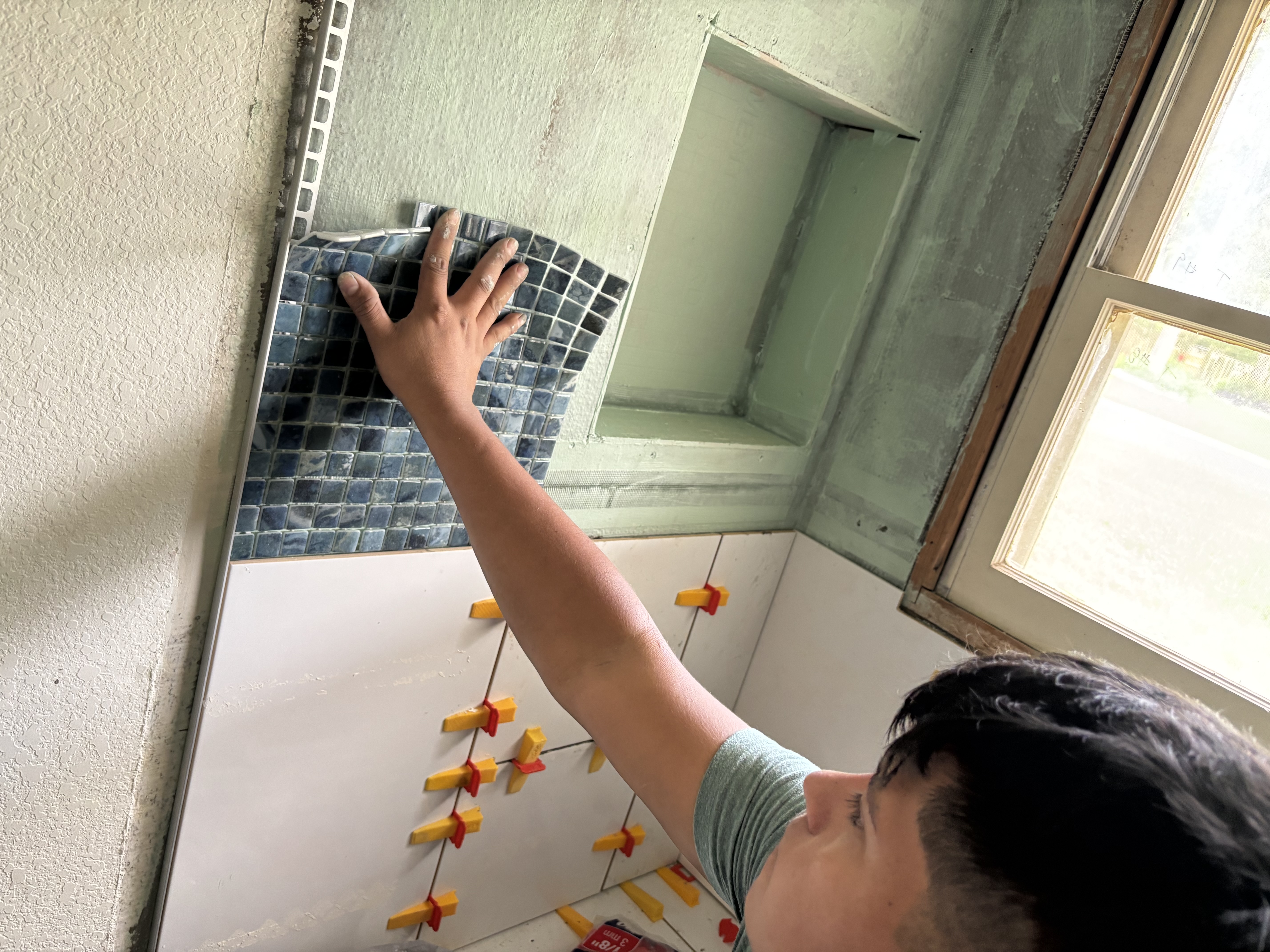 A Gutierrez Flooring installer pressing blue glass mosaic tile into a custom shower niche.
