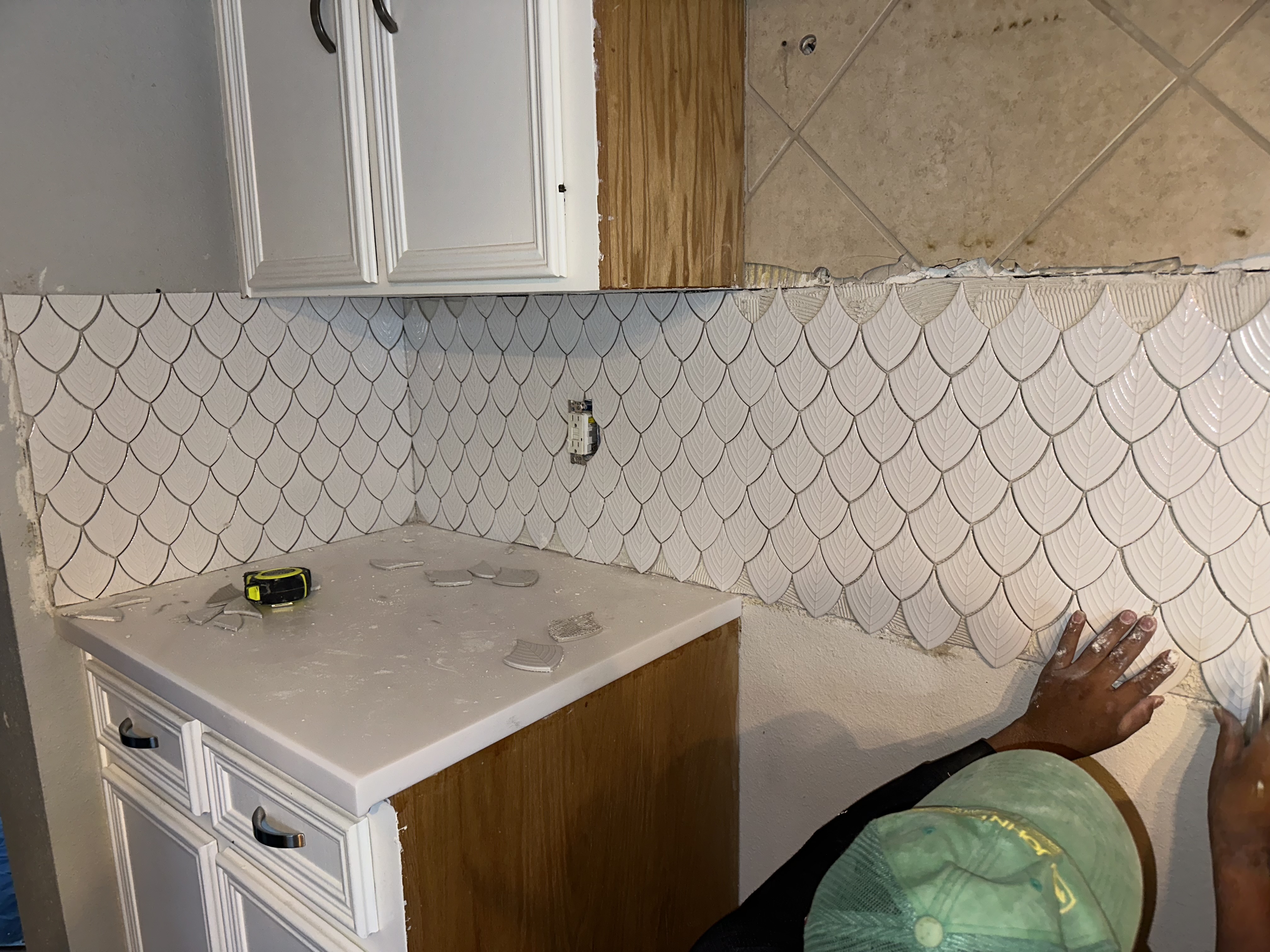 White fish-scale tile backsplash being hand-set in a kitchen, worked neatly around the outlet and countertop